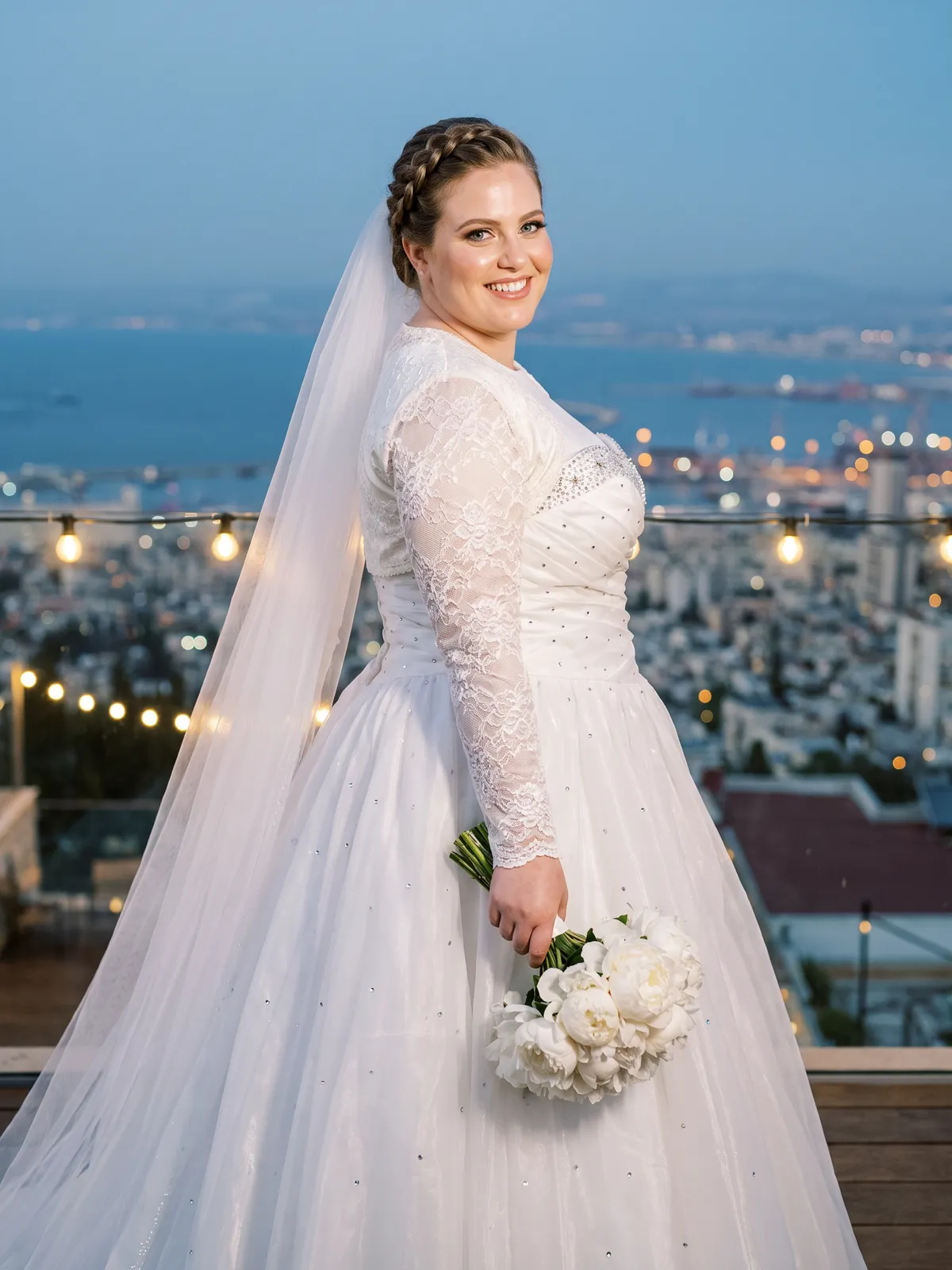 שמלת כלה צנועה עם מחוך סאטן רקום חרוזים וחצאית אורגנזה מלאה, עם בולרו תחרה אטום בשרוולים ארוכים, במרפסת על הכרמל מעל מפרץ חיפה בשעת דמדומים