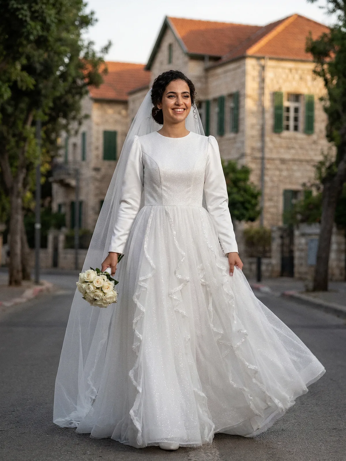 שמלת כלה צנועה עם חלק עליון מבד מנצנץ, שרוולי סאטן ארוכים וחצאית טול עם מפלי מלמלה, בשדרה במושבה הגרמנית בחיפה