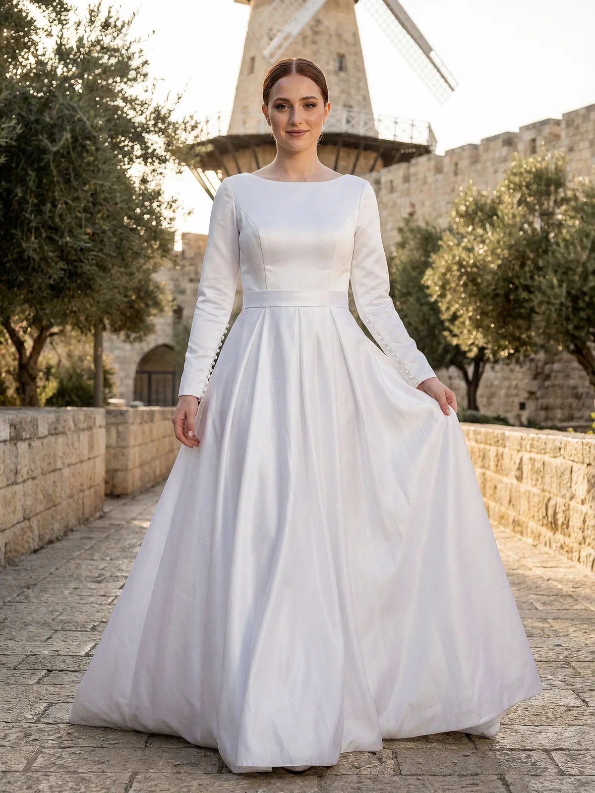 שמלת כלה צנועה מסאטן עם צווארון סגור, חגורת סאטן רחבה ושרוולים ארוכים עם כפתורי פנינים, ליד טחנת הרוח בימין משה בירושלים