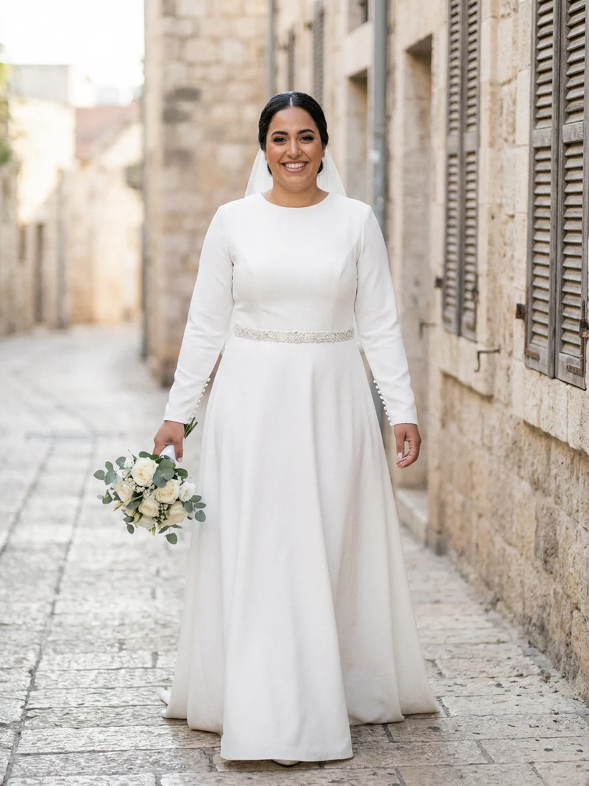 כלה בשמלת כלה צנועה מקרפ בגזרת A עם צווארון סגור, שרוולים ארוכים וחגורת פנינים, ברחוב היסטורי בראשון לציון