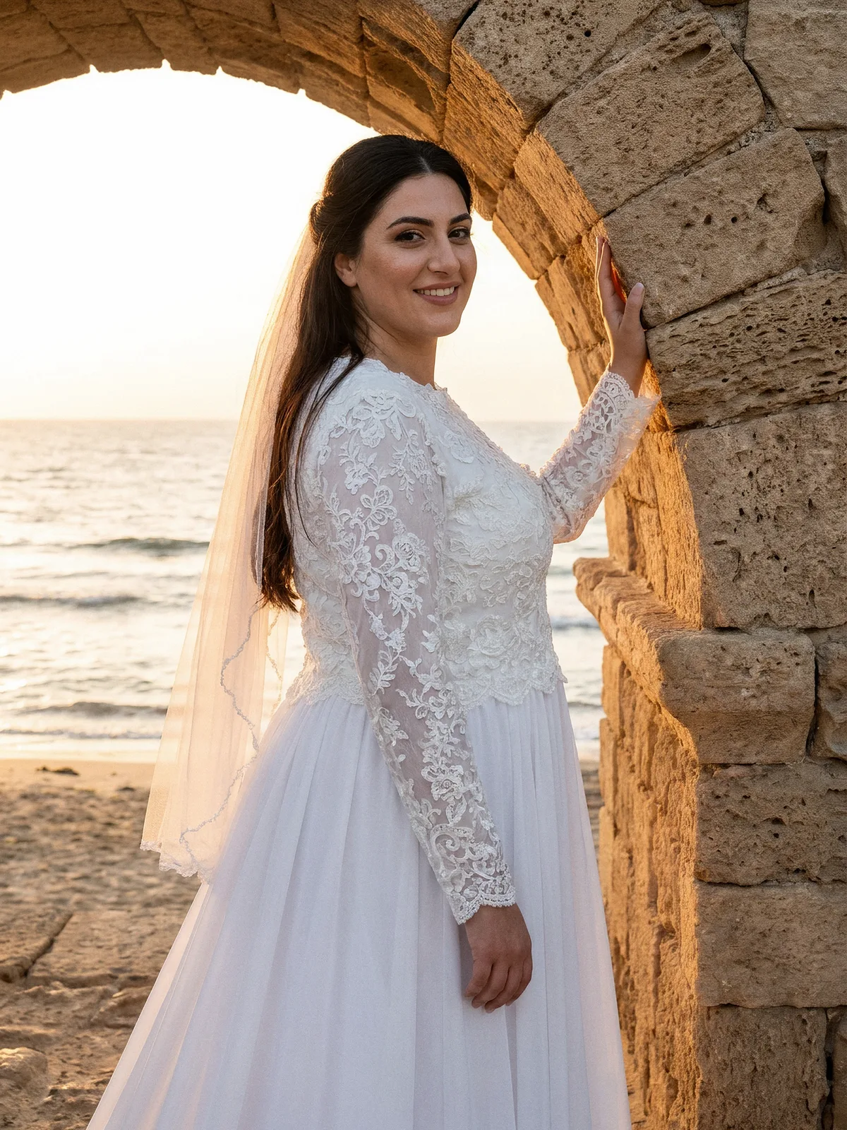 כלה בשמלת כלה צנועה עם חלק עליון מתחרה, שרוולי תחרה ארוכים וחצאית שיפון זורמת, ליד אמת המים העתיקה בקיסריה בשקיעה